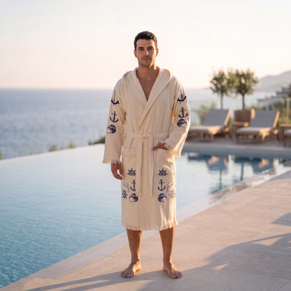 A man wearing a cream-colored hooded peshtemal bathrobe with navy blue anchor and nautical motifs, standing on a poolside patio with a scenic ocean view in the background.