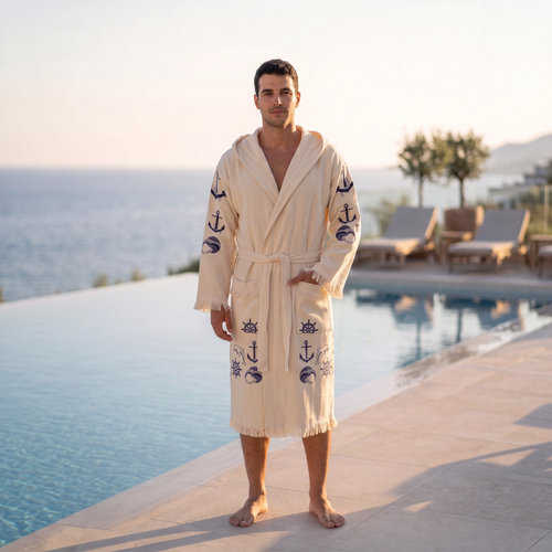 A man wearing a cream-colored hooded peshtemal bathrobe with navy blue anchor and nautical motifs, standing on a poolside patio with a scenic ocean view in the background.