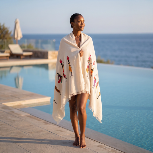 A woman stands barefoot by an infinity pool wearing a cream-colored fringed peshtemal with colorful illustrated figures, with a blurred ocean and poolside setting in the background.