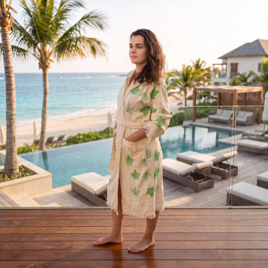 A woman stands on a wooden deck overlooking an oceanfront resort pool and lounge area, wearing a cream peshtemal bathrobe with green leaf embroidery.