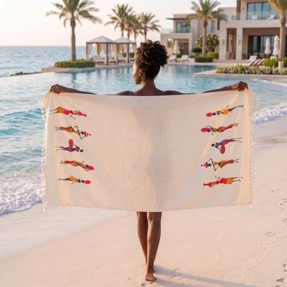A woman stands on a sandy beach facing the ocean, holding up a large cream-colored peshtemal with colorful illustrations of stylized figures and white fringe on the ends.